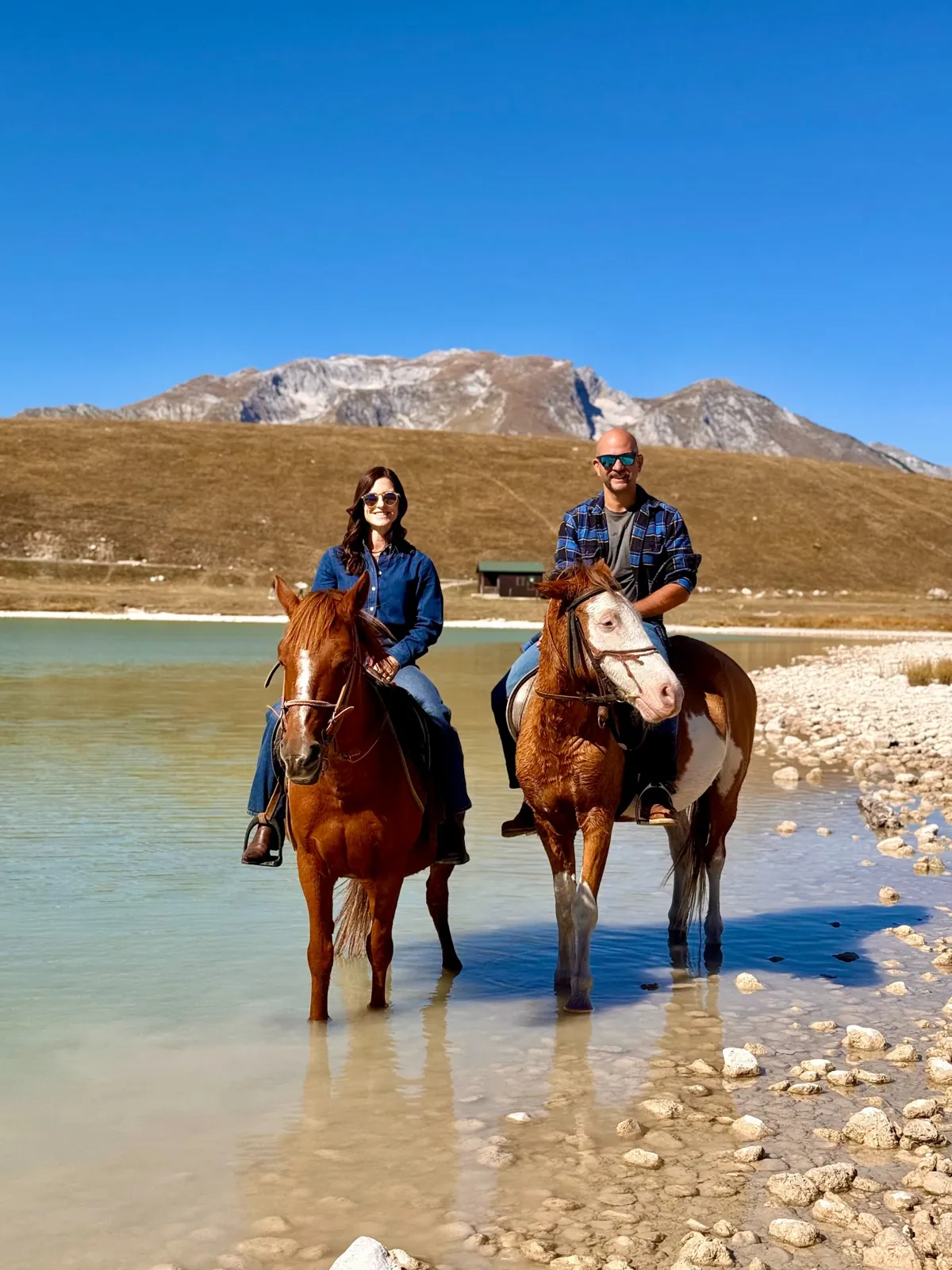 Two guests on horseback beside a turquoise lake with Durmitor mountains behind them